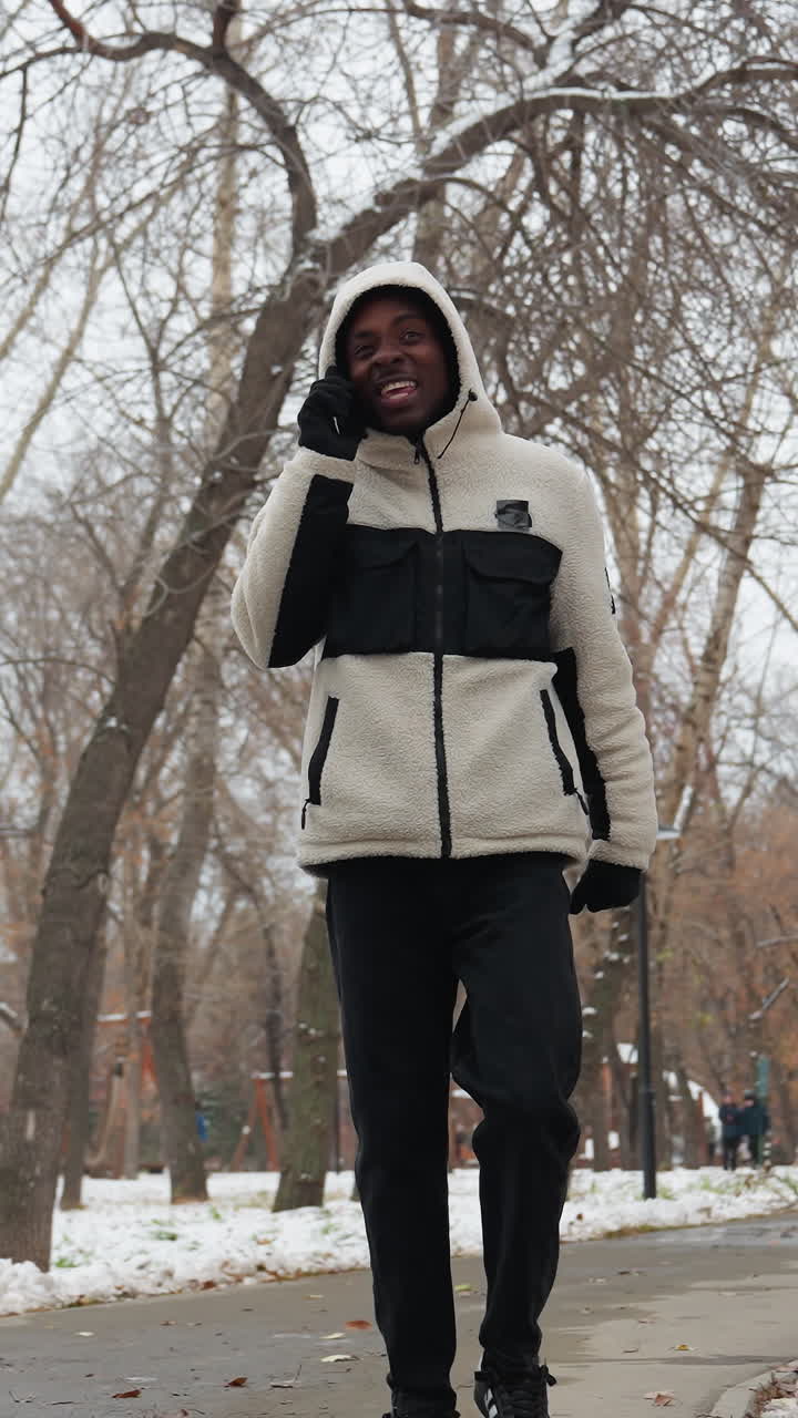 hombre africano hablando por teléfono con alguien mientras paseaba por el parque durante el invierno, lleva una sudadera blanca con capucha y guantes, con árboles, nieve y edificios borrosos en el fondo