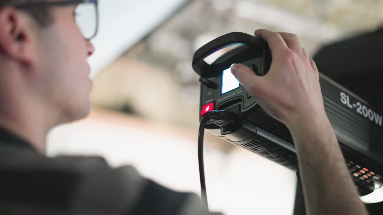 Camera technician operating LED panel control, pressing power switch and adjusting brightness knob on mounted studio light with cables visible against blurred background during broadcast production