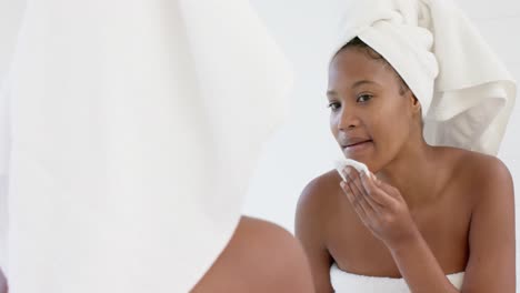 feliz mujer biracial con toalla en la cabeza mirando en el espejo lavándose la cara, cámara lenta