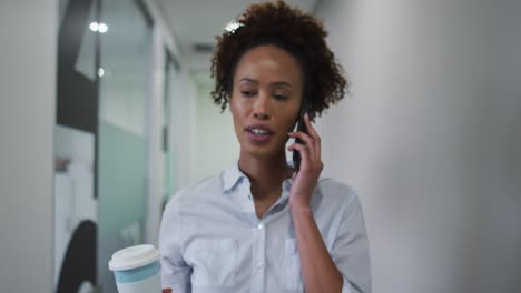 Mixed-race-businesswoman-talking-on-smartphone-walking-with-coffee-in-office-corridor