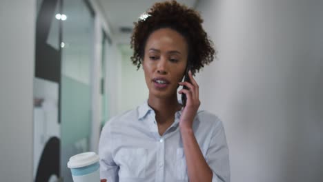 mixed race businesswoman talking on smartphone walking with coffee in office corridor