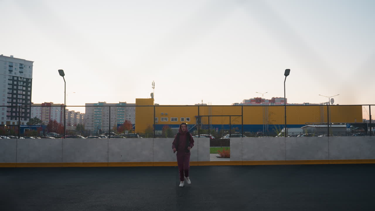 Jogger running toward chain link fence on dusk lit urban court under pastel sky framed by high rise apartments and yellow industrial building, metal mesh barrier, evening exercise scene