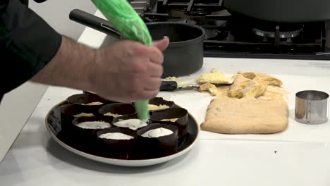 A chef skillfully pipes sweet cream into chocolate molds while working in a contemporary kitchen. Crumbled cake pieces are visible on the counter, indicating preparation for a delicious dessert.