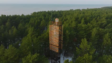 vista aérea de pájaro del faro de bernati rodeado por un exuberante bosque de pinos verdes con nieve ligera, bosque nórdico, costa del mar báltico, día soleado de invierno, letonia, amplio tiro de drones en órbita