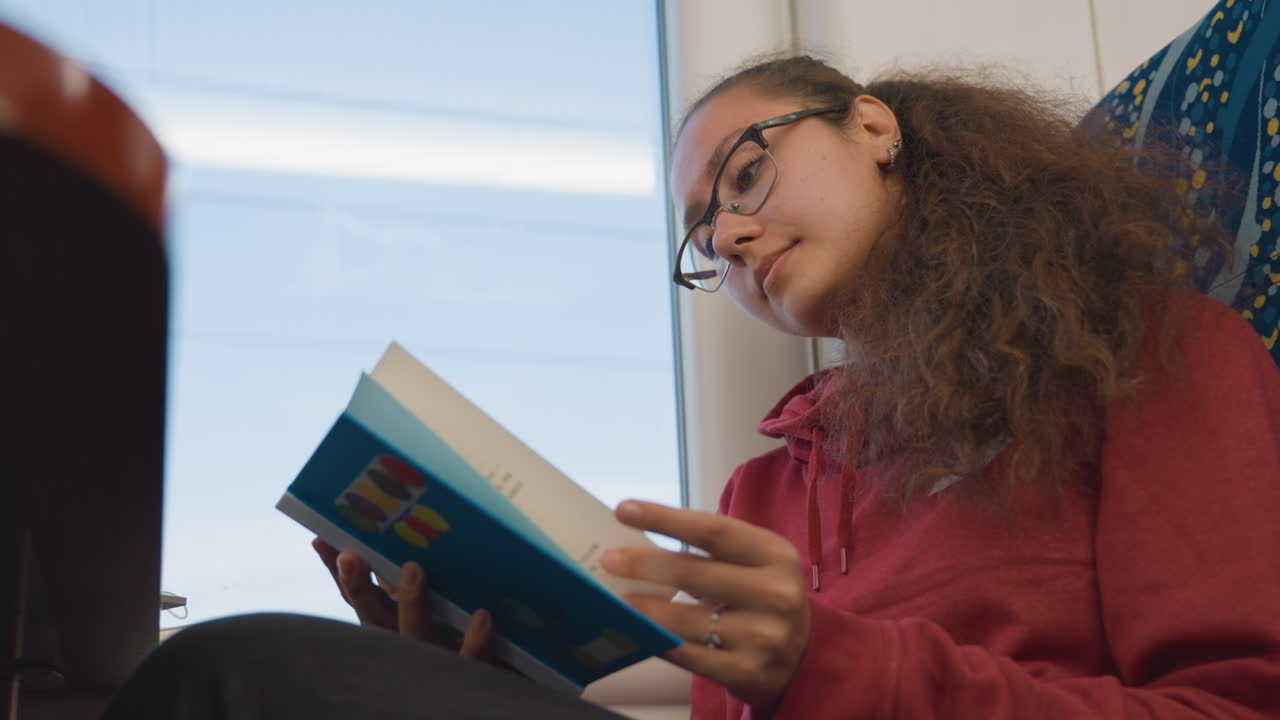 Voyageur du soir plongé dans un livre. Passager étudiant avec des lunettes lors de son trajet nocturne. Voyageur concentré sur un livre en parcourant le paysage urbain la nuit, sous un éclairage intérieur doux.