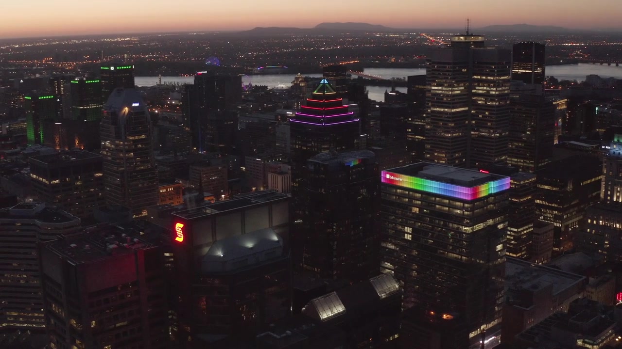 Neon-trimmed Montréal high-rises light up at blue hour as the camera slowly flies past the downtown core toward the river.