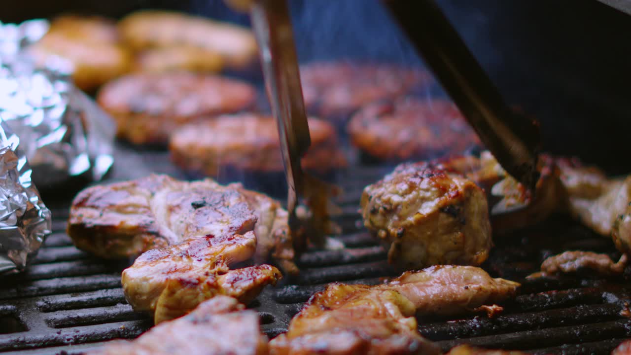 chef usando pinzas para girar la chuleta de cerdo en la barbacoa de fumar caliente con chuletas de cordero, hamburguesas y salchichas en cámara lenta 4k