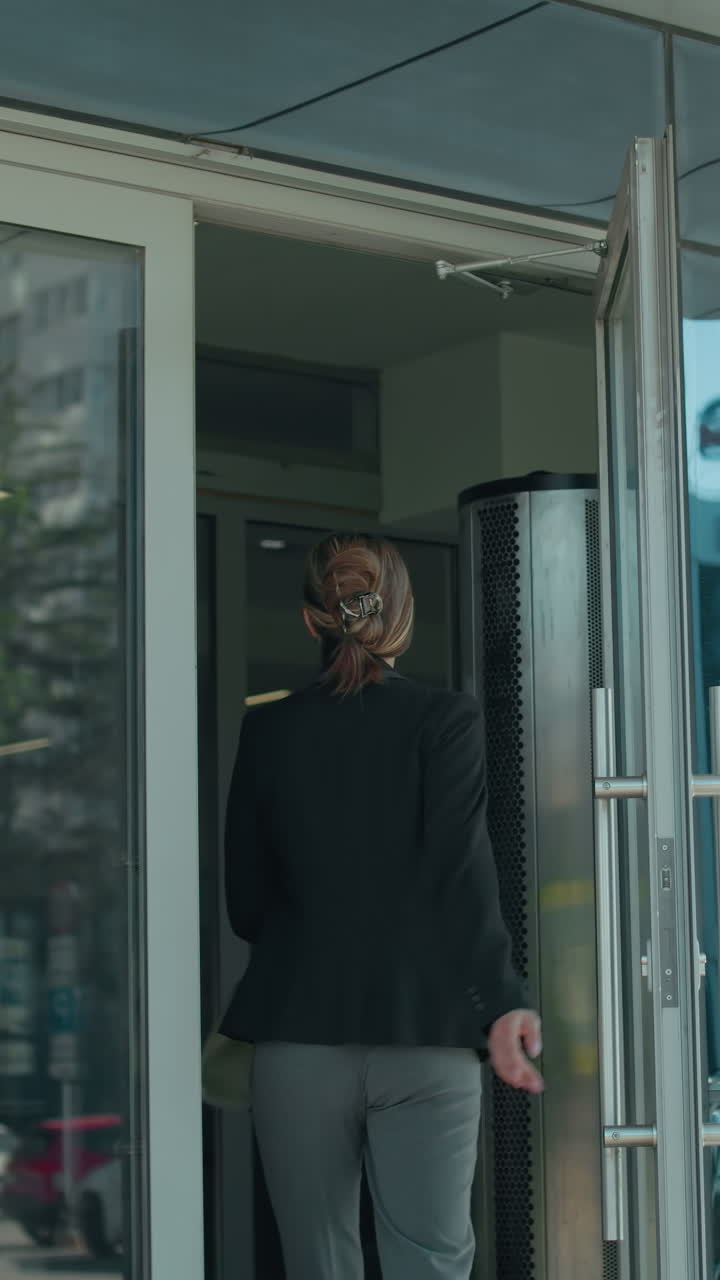 Office worker walks into office building carrying handbag, dressed professionally in black jacket and gray trousers, with glass door reflecting urban street, parked cars, and cityscape on sunny day