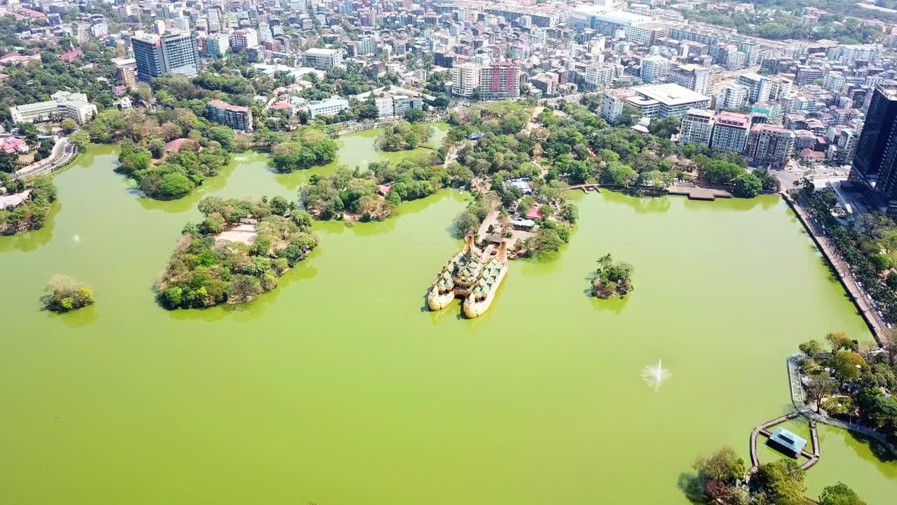 Aerial view of Kandawgyi Lake and Karaweik Palace in Yangon, Myanmar, showcasing the vibrant green water, lush greenery of the park, and the bustling cityscape in the background on a sunny day