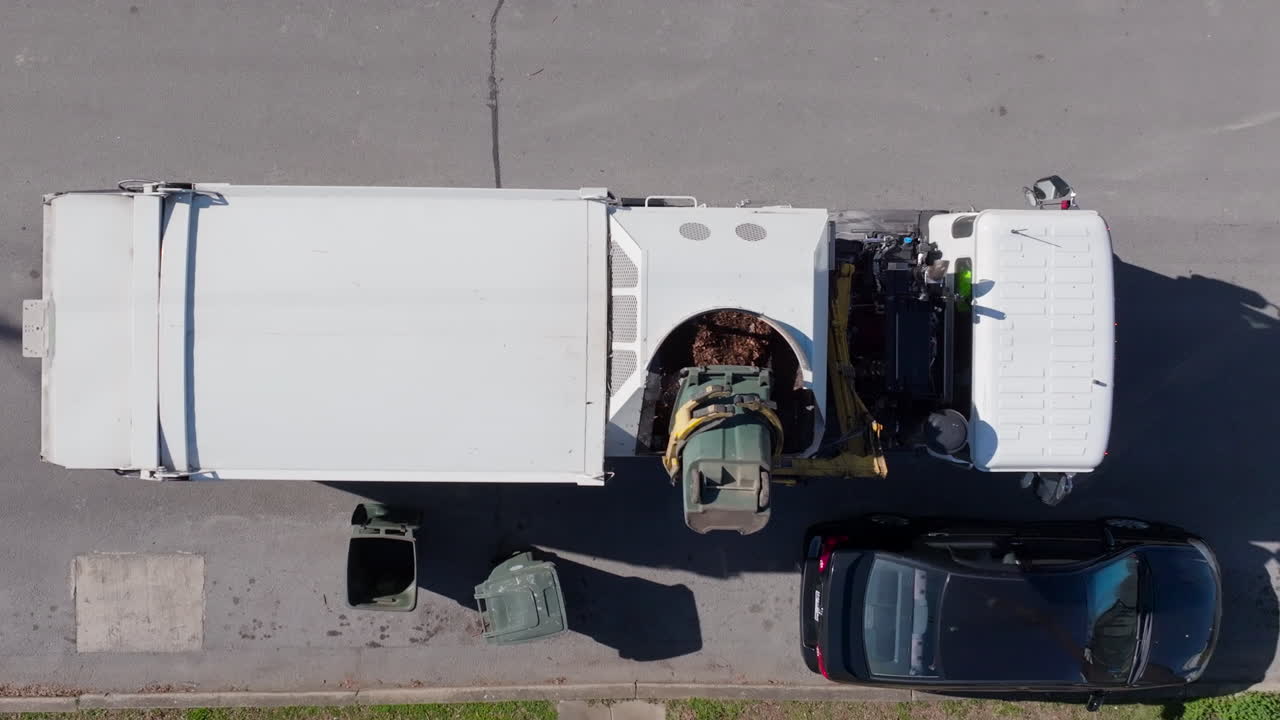 Top down aerial footage of trash truck using a mechanical arm to pick up a trash can for disposal.