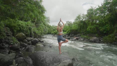 behind blond woman practicing yoga beside tropical river, tree pose, bali