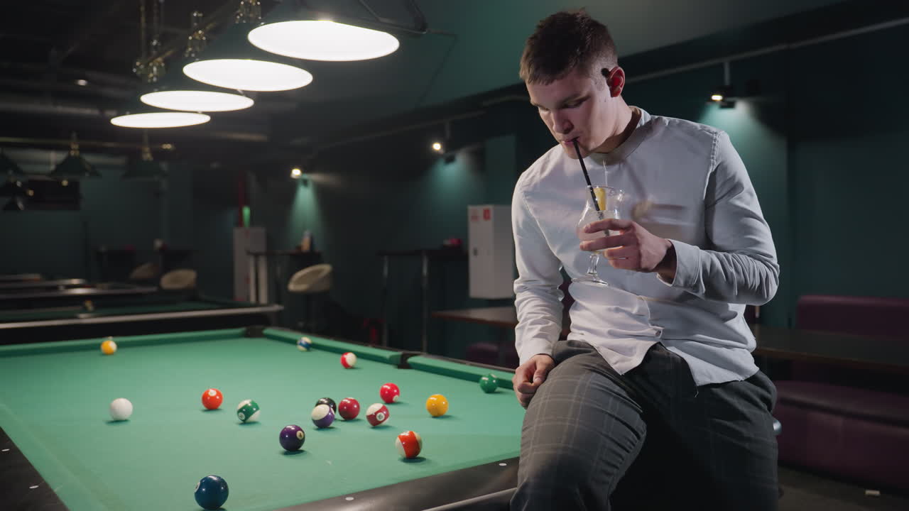 jugador de billar con camisa blanca sentado en una mesa de billar verde, bebiendo una bebida de limón con paja negra en una sala de billar débilmente iluminada. luces superiores, bolas de billar de colores esparcidas en la mesa