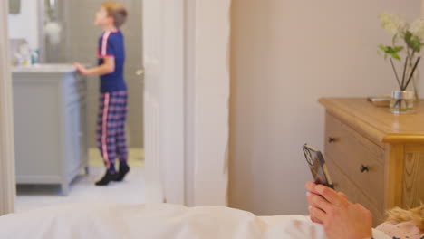 Mother-Looking-At-Mobile-Phone-In-Bed-As-Son-Brushes-Teeth-Looking-In-Mirror-In-Bathroom-At-Home