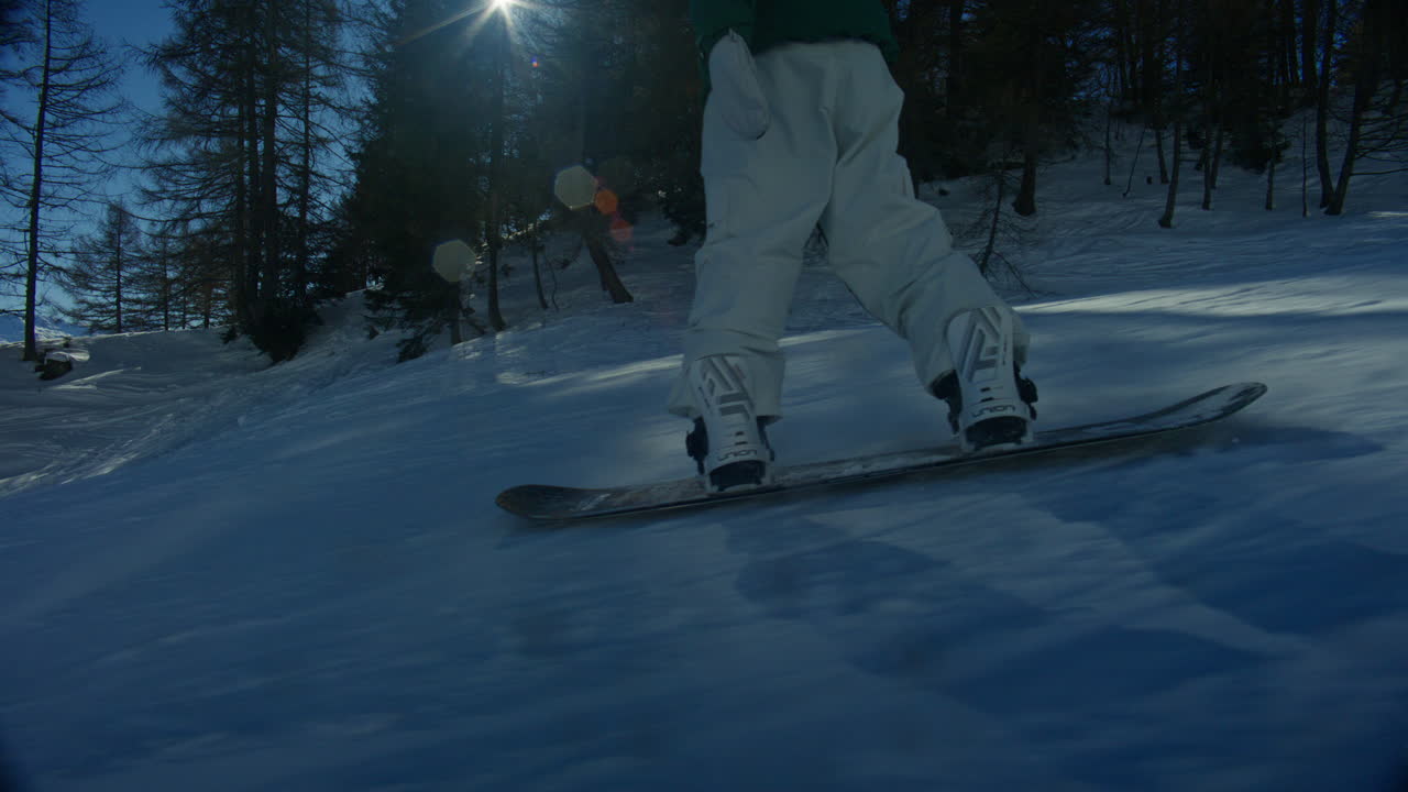 Ujęcie z kamery śledzącej snowboardzistę sunącego i rzeźbiącego na przygotowanym stoku w Anzère w słoneczny dzień. Drzewa otaczają trasę, gdy światło słoneczne znika i pojawia się, tworząc naturalne flary obiektywu i kinowy efekt.