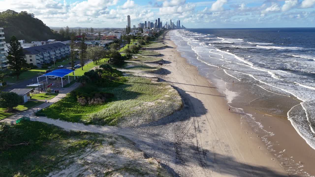 Drone footage captures Gold Coast's sandy shoreline and ocean waves under clear skies, showcasing coastal beauty and urban skyline