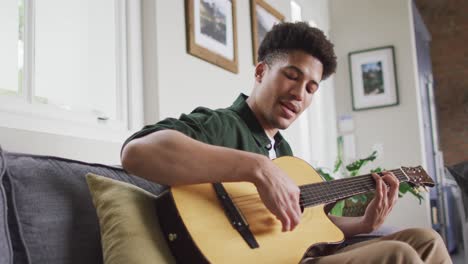 happy biracial man sitting on sofa in living room playing guitar