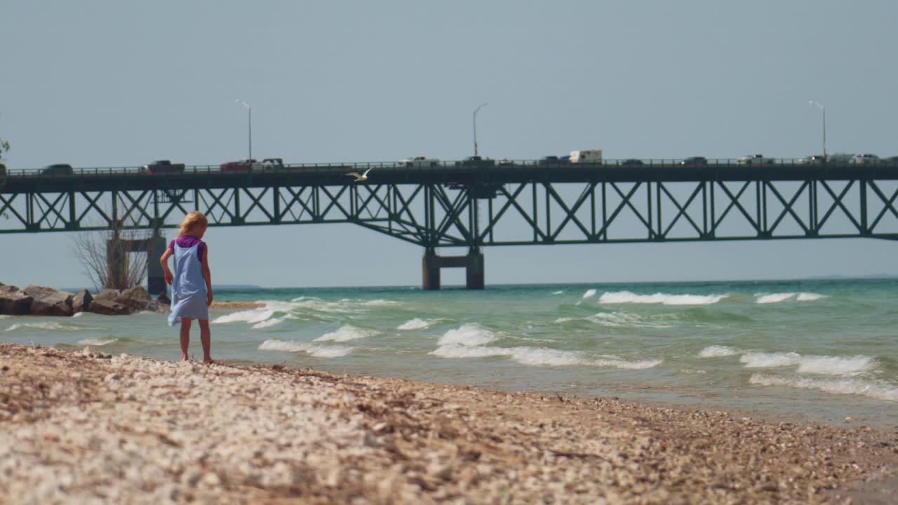Wide Shot of Cute Little Girl Throwing Rocks into the Sea with Large Bridge in the Background