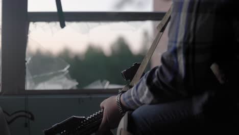 close up of rifle and sling held by mercenary solider sitting next to broken glass window in abandoned building on guard duty as sun rises