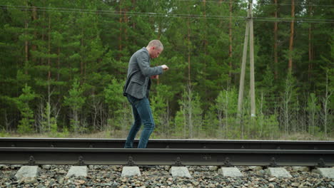 a man wearing a grey suit jacket and blue jeans practicing taekwondo along railway tracks in a forested area, with greenery and electric poles in the background