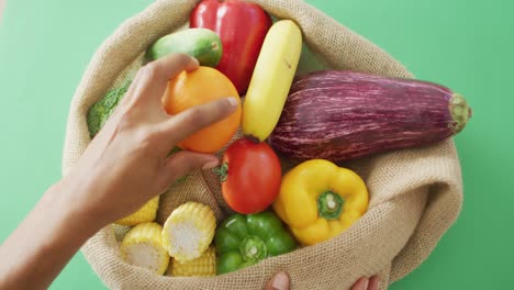 video of biracial woman arranging fresh fruit and vegetables in rustic bag on green background