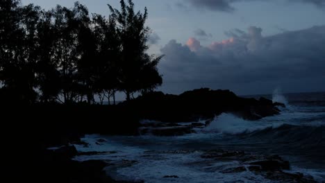 powerful ocean waves crash into rugged black volcanic rocks under a dark twilight sky, with silhouetted trees enhancing the dramatic coastal atmosphere.