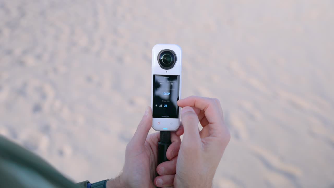 Mãos seguram uma câmera de ação 360 em um bastão de selfie ao ar livre, verificando a visualização prévia e as configurações na tela touchscreen