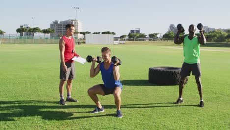 Grupo-Diverso-De-Dos-Hombres-En-Forma-Y-Un-Entrenador-Masculino-Haciendo-Ejercicio-Al-Aire-Libre-En-Cuclillas-Y-Levantando-Pesas