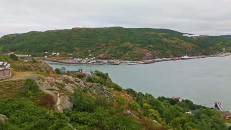 A drone hovers above Brigus South harbour, capturing the wharf with a tall mast, calm waters, fishing boats, and a colorful hillside community along Newfoundland coast