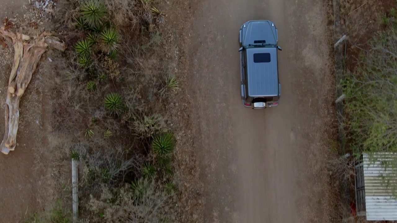 Vista aerea dall'alto con drone di un veicolo utilitario sportivo che guida lungo una strada sterrata rurale in Messico, caratterizzata da vegetazione secca, un sentiero non asfaltato e un paesaggio di campagna