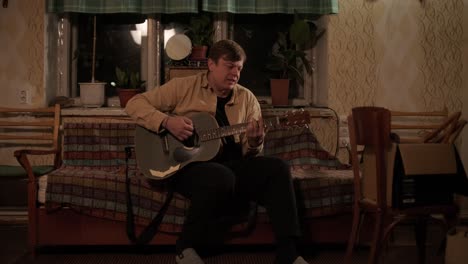 pretty young man playing guitar while sitting on sofa in dark living room