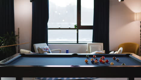 a person prepares to take a shot at a pool table set up in a comfortable room with natural light. surrounding chairs and plants enhance the inviting atmosphere.