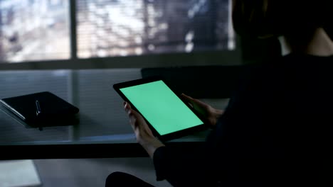 sobre la vista del hombro de la mujer de negocios sentada en su escritorio y usando una tableta con pantalla verde. la gran ciudad se ve por la ventana.