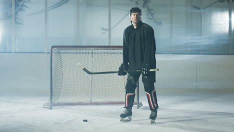 Porträt-Eines-Konzentrierten-Männlichen-Hockeyspielers-In-Uniform-Und-Helm,-Der-Mit-Schläger-Und-Puck-Auf-Der-Eisarena-Steht,-Während-Er-In-Die-Kamera-Blickt