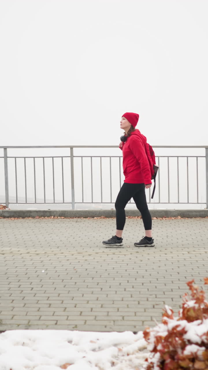 Side view of athlete in red sweater with bag over shoulder walking near iron railing on snow dusted ground with scattered leaves surrounded by foggy atmosphere creating calm serene setting