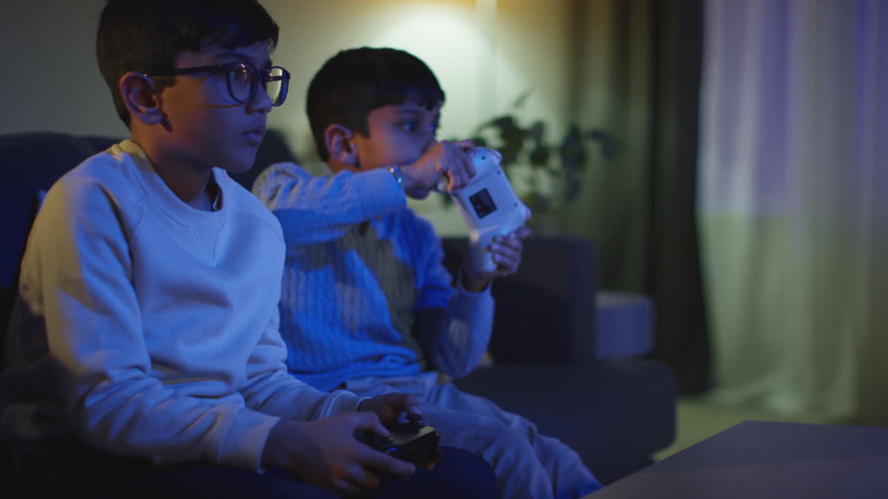 Two Young Boys Sitting On Sofa At Home Playing With Computer Games Console On TV Holding Controllers Late At Night 4