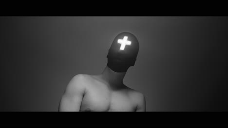 masked and hooded man with cross of light glowing on his face looks in the camera