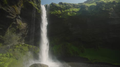 kvernufoss waterfall in iceland, sunny green rocks in the foreground and small figures hiking underneath