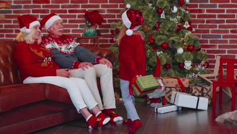 Grandchild-girl-gifting-Christmas-Xmas-present-box-to-amazed-happy-grandparents-at-home,-celebrating