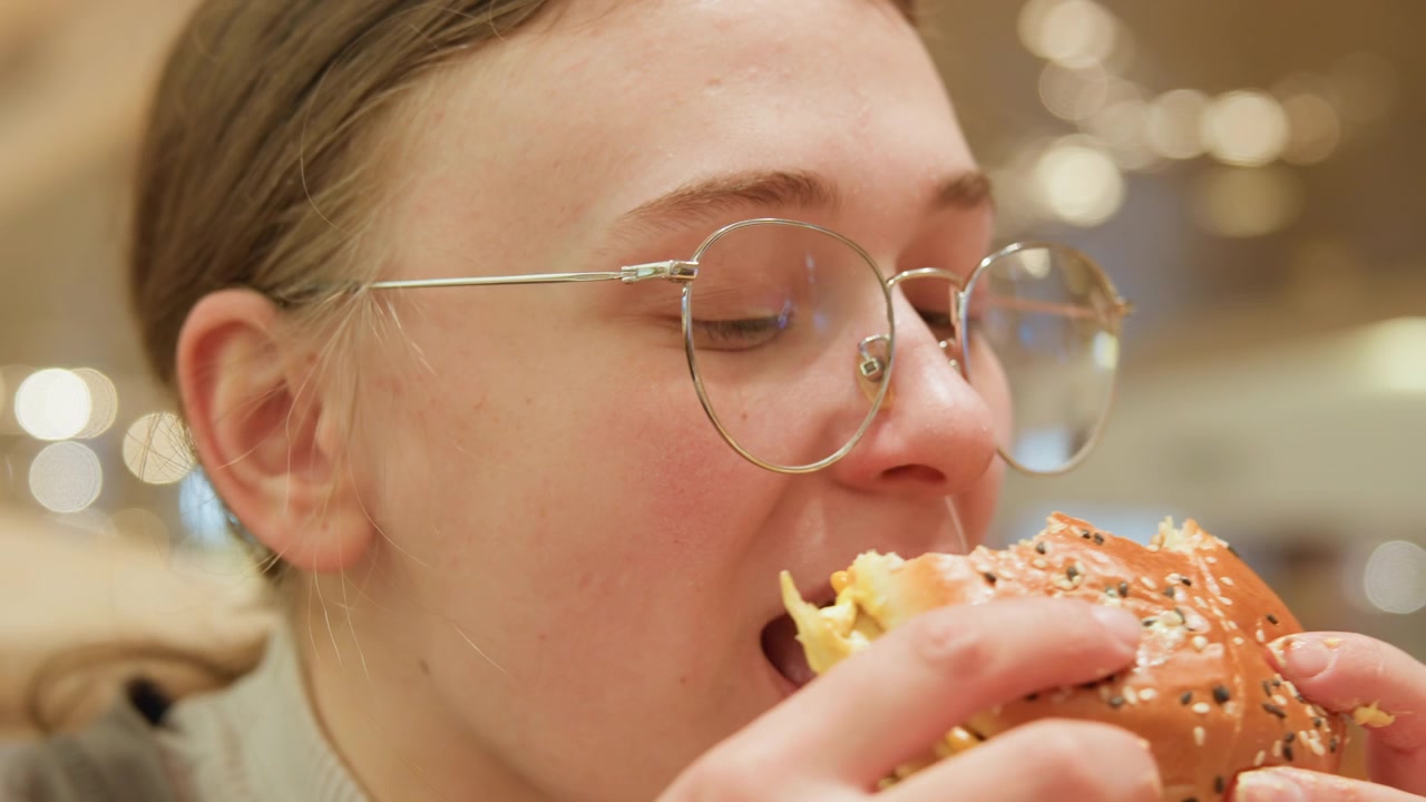 White college student savoring comfort burger with glasses gentle closedeyes expression during slow bite, seeded bun and melted cheese visible, ambient mall lights and relaxed dining mood