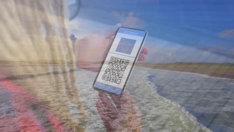 Composition-of-woman-holding-a-smartphone-with-qr-code-on-screen-against-aerial-view-of-the-beach