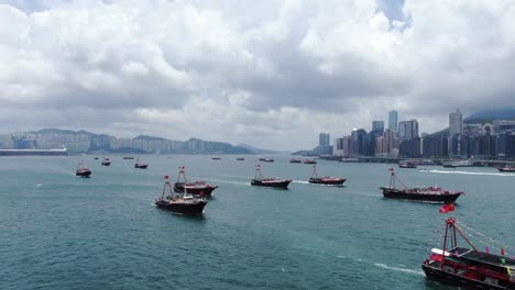 Convoy-of-local-Fishing-boats-causing-in-Hong-Kong-Victoria-bay,-with-city-skyline-in-the-horizon,-Aerial-view