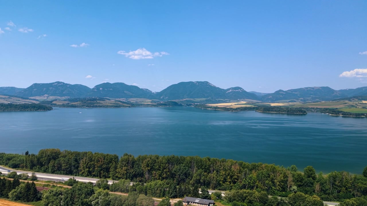 スロバキアの森と野原に囲まれた山の湖。道端の木々の向こうに広がる広い谷を青い水が満たし、地平線には重なり合う山並みが続く