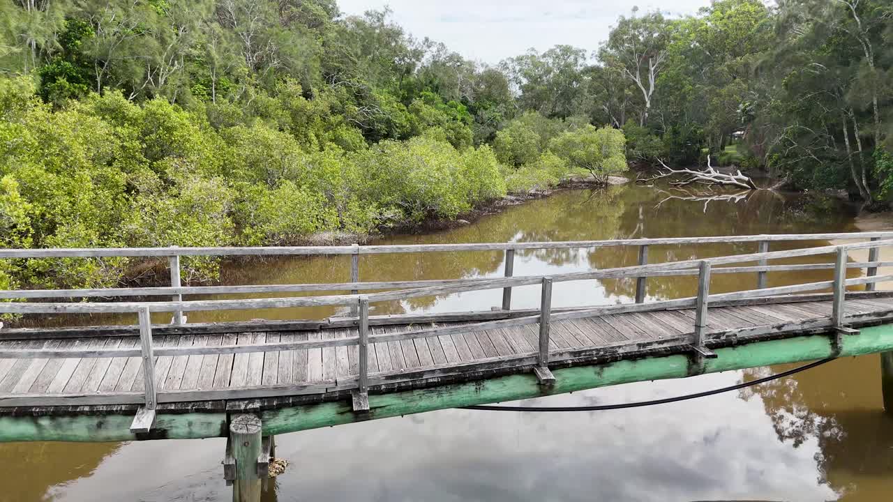 Drone glides over rustic wooden bridge spanning creek, lush greenery, calm water, soft daylight