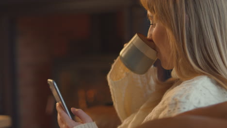 Close-Up-Of-Woman-At-Home-In-Lounge-With-Cosy-Fire-And-Hot-Drink-Using-Mobile-Phone