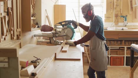 african american male carpenter cutting wooden plank with electric chop saw in a carpentry shop