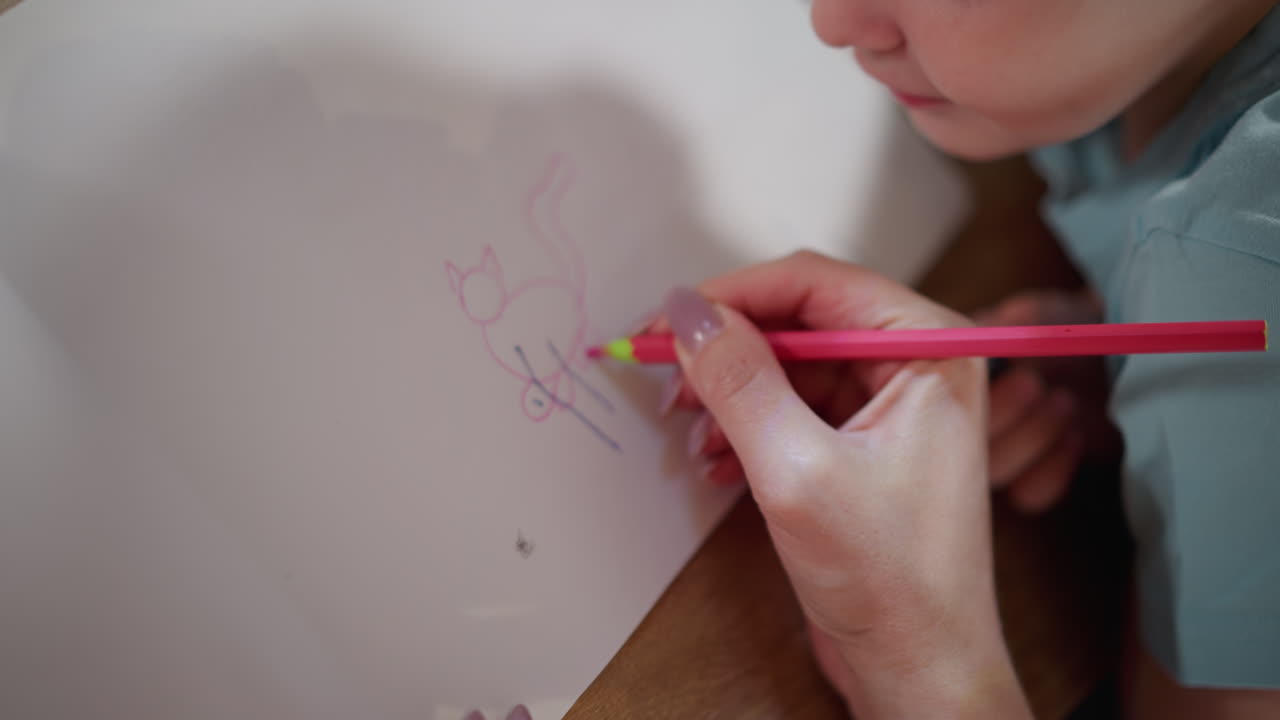 Closeup hand using pink pencil sketching simple cat figure on white paper while child holds black pen, focusing on drawing practice, creativity, parent teaching child basics of art with patience