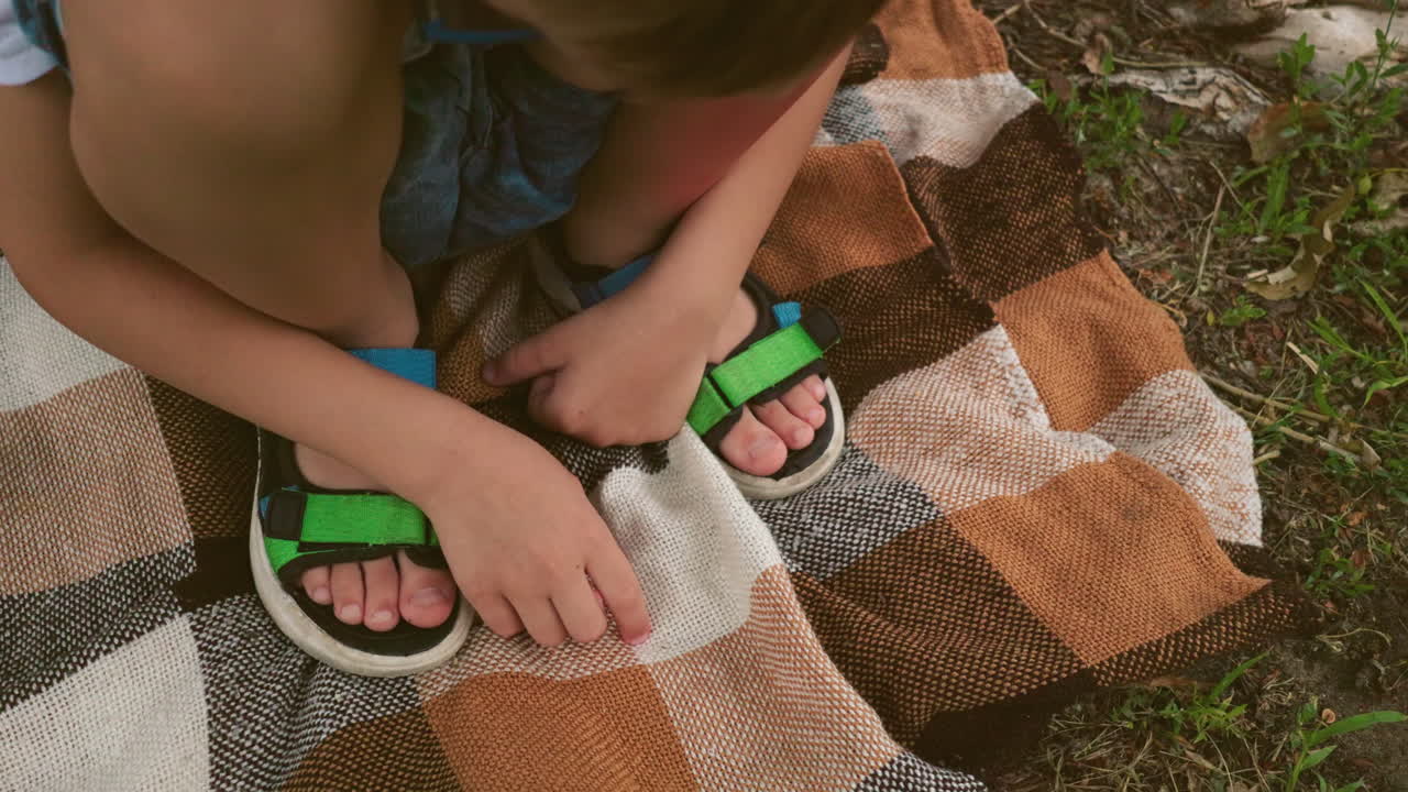 Close-up of a child squatting on a checkered blanket with one hand touching it and the other hand resting on his leg, wearing a green sandal