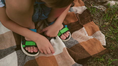 close-up of a child squatting on a checkered blanket with one hand touching it and the other hand resting on his leg, wearing a green sandal