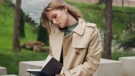 caucasian female student talking on the phone with a notebook in the park.