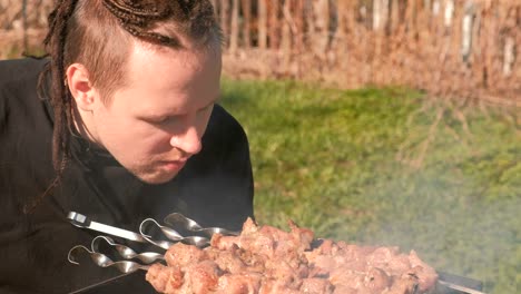 young men with dreads on hairs cooks shashlik meat on top of charcoal grill on backyard.